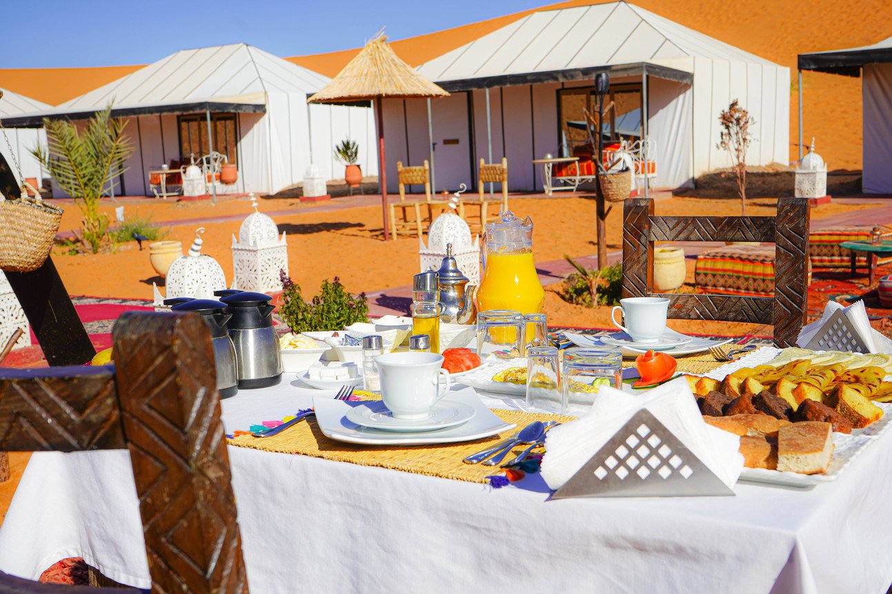 Breakfast table with bread, pastries, orange juice and hot drinks beside white tents and orange dunes in Morocco