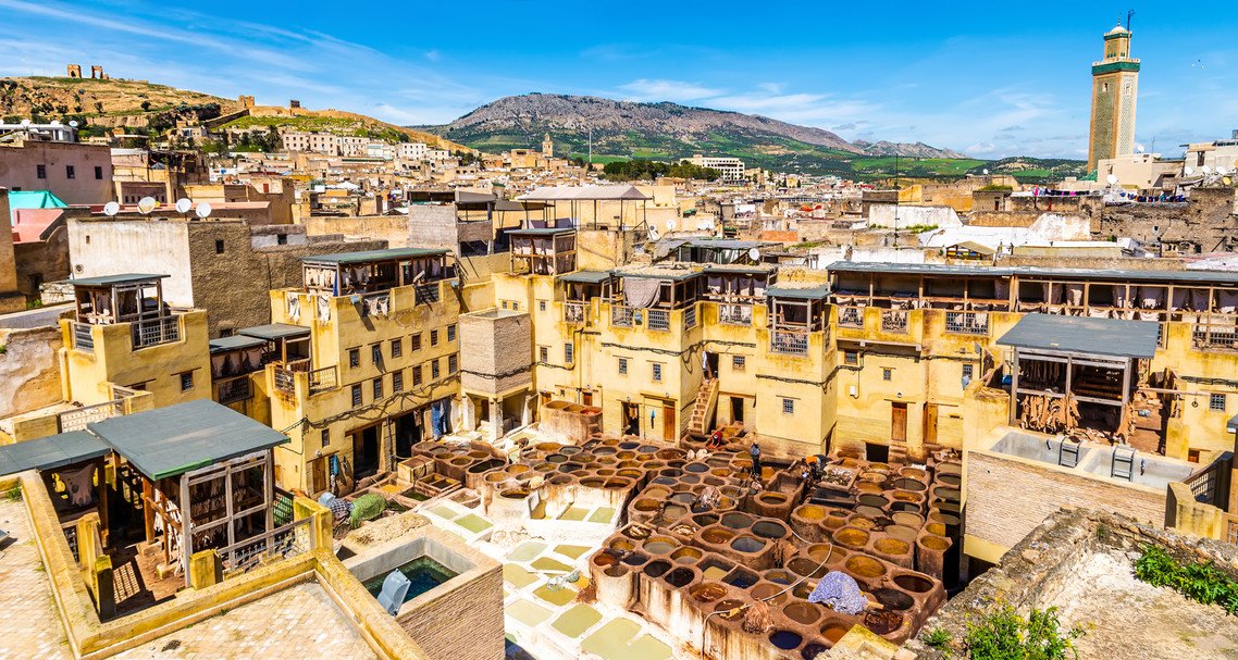 Chouara Tanneries dye vats surrounded by historic Fes medina buildings, with hills and a minaret beyond