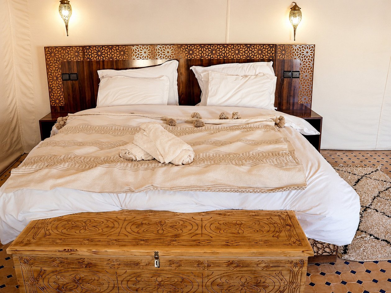 Large double bed inside a Moroccan desert camp tent with two warm lamps, a carved headboard, cream linens and chest