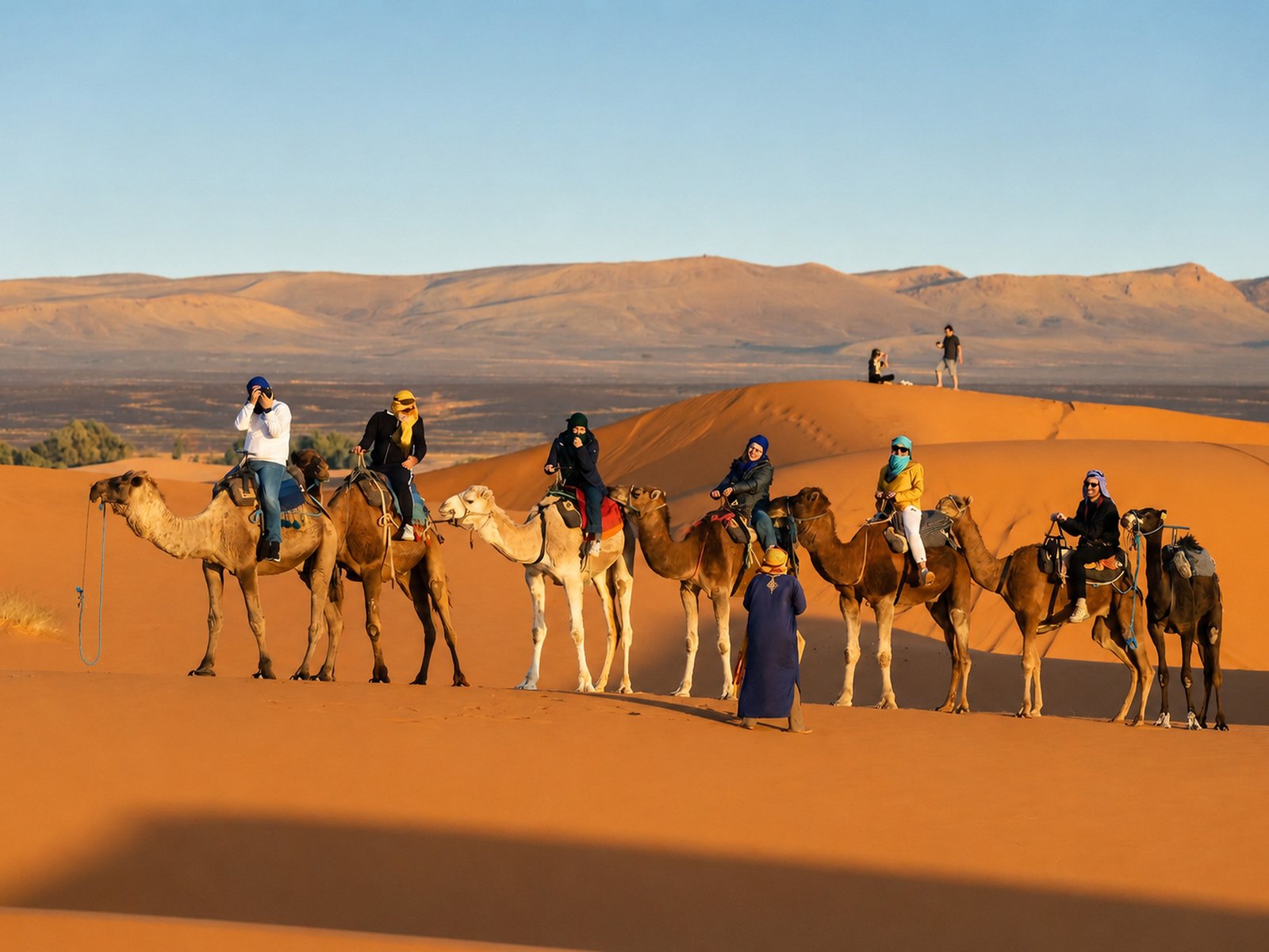 Travelers riding a close line of camels with a guide walking beside them across orange desert dunes in Morocco