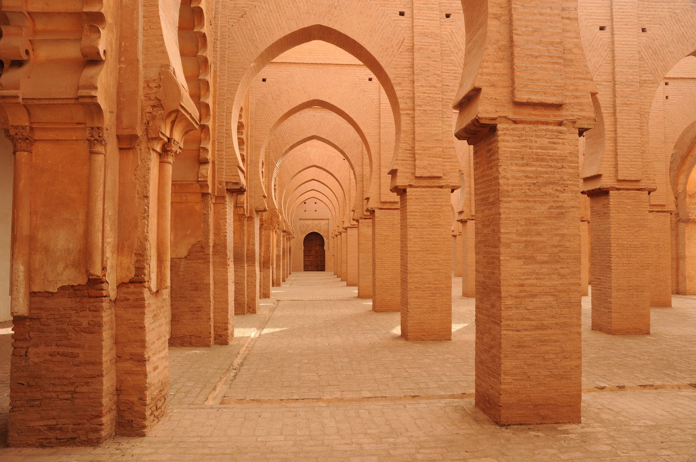 Rows of warm brick columns and pointed arches forming a symmetrical historic Moroccan mosque interior
