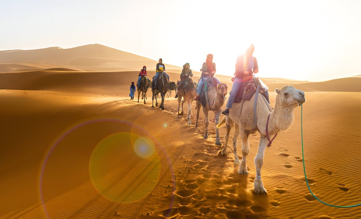 Travelers riding camels in a caravan across golden Moroccan Sahara dunes near sunset