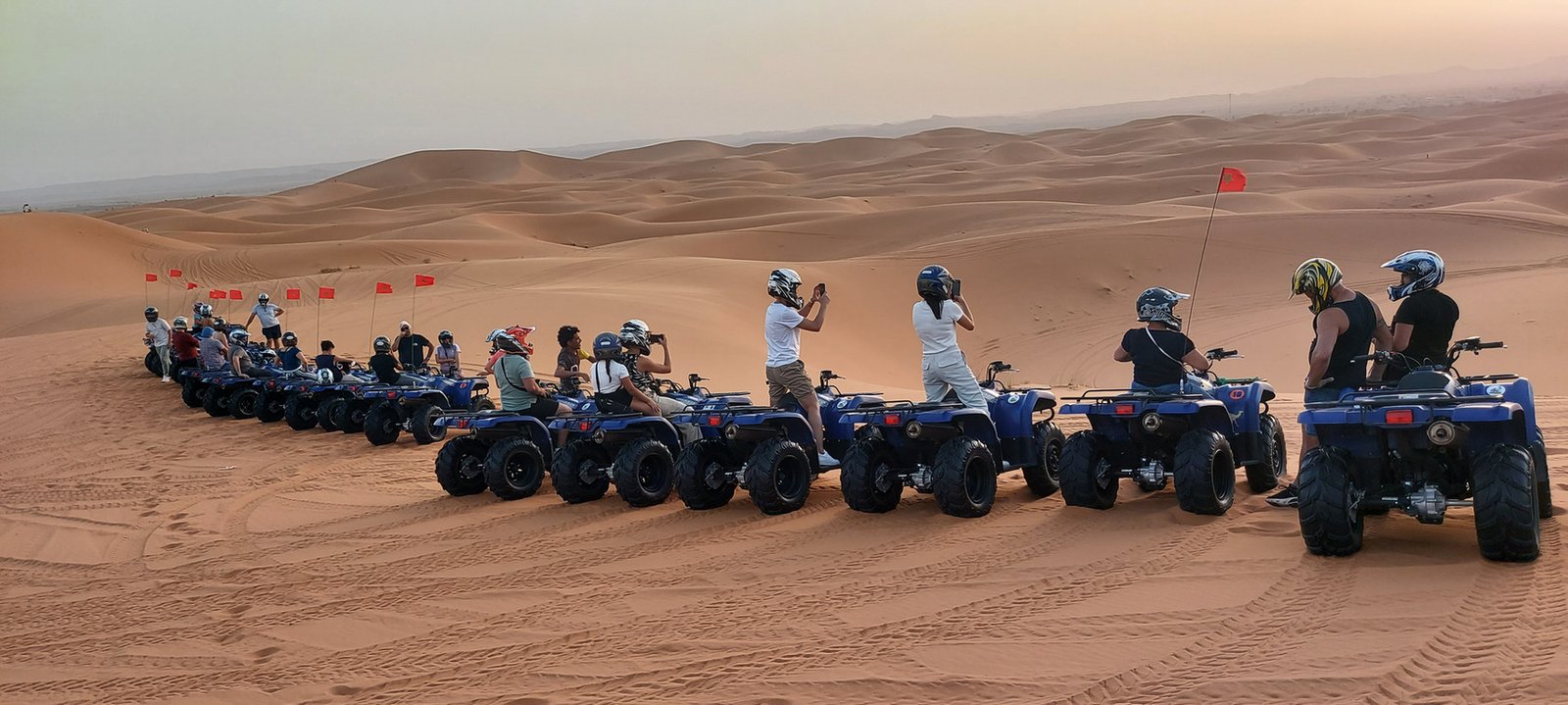 Travelers with blue quad bikes lined up on a desert dune marked by Moroccan flags