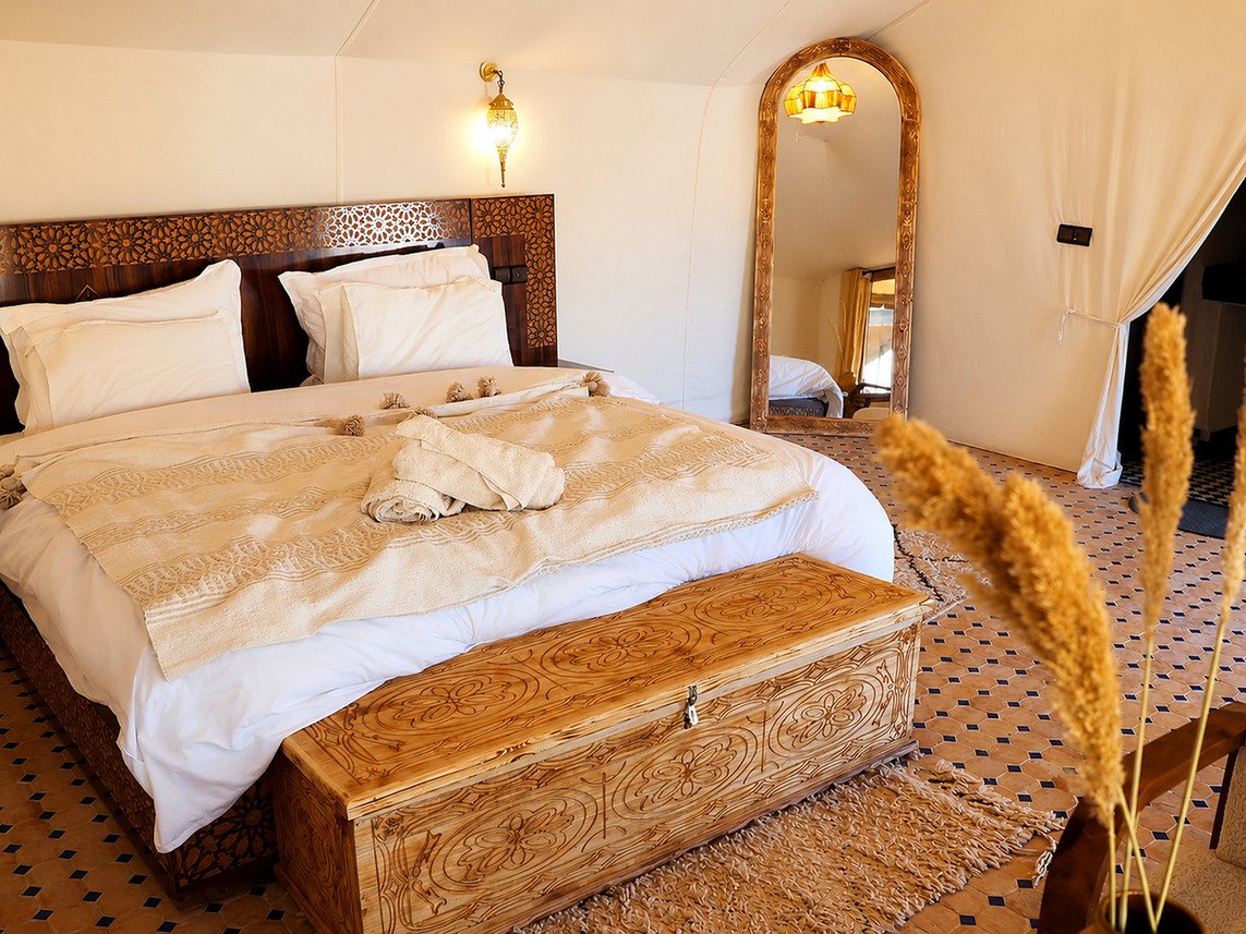 Wide Moroccan desert camp tent with a large double bed, carved chest, floor mirror, woven rug and warm wall lighting