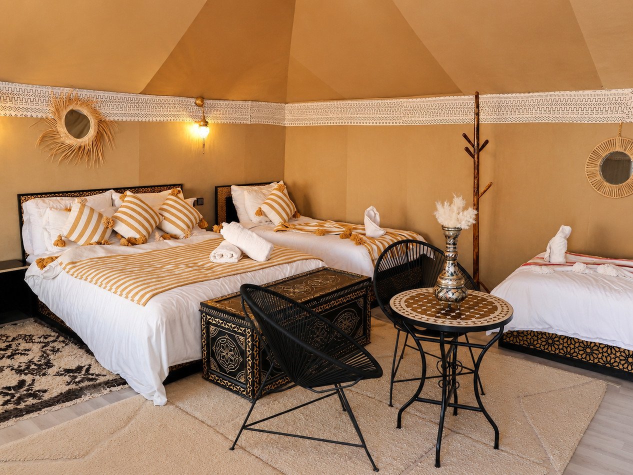 Large tented room with one double bed, two single beds, gold-striped textiles, carved chests and a small seating table