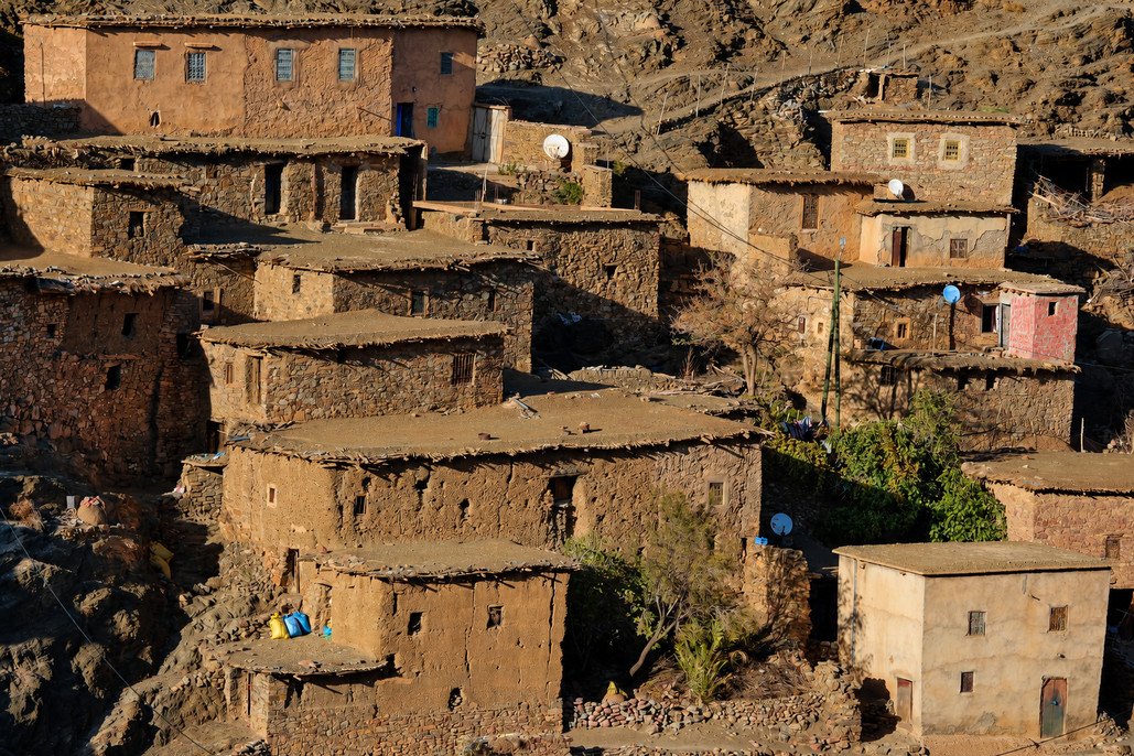 Stone and earthen village houses with flat roofs clustered across a rocky mountainside in rural Morocco