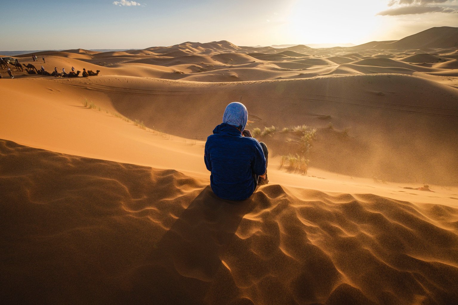 Traveler seated on a sand dune overlooking golden Moroccan Sahara ridges at sunset, with camels in the distance